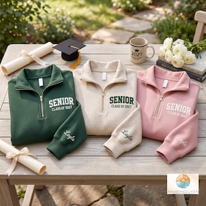 Op de afbeelding: Drie kwart-rits sweatshirts in donkergroen, crème en lichtroze, elk met de tekst SENIOR CLASS OF 2027 op de borst en de naam Layla geborduurd op de mouw. De sweatshirts zijn gerangschikt met een afstudeerhoed en een diploma.