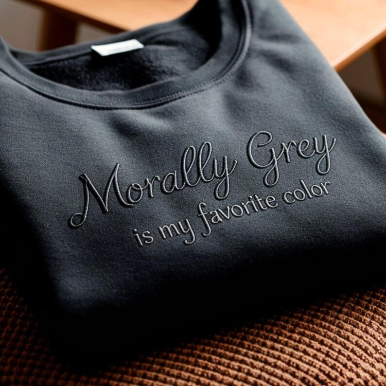 May include: A dark gray sweatshirt with the embroidered text "Morally Grey is my favorite color." The sweatshirt is folded and resting on a brown surface. The text is in a cursive font.