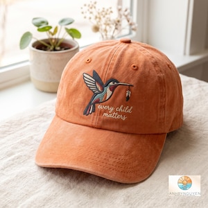 Puede incluir: Una gorra de béisbol naranja con un bordado de un colibrí que lleva una pluma en el pico. Debajo del ave, el texto "every child matters" está bordado en hilo blanco. La gorra tiene una visera curva y una corona estructurada con ojales.