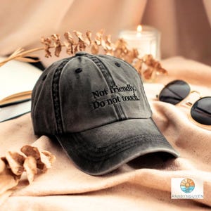 Peut inclure: Une casquette de baseball gris anthracite avec une finition délavée et usée. L'avant de la casquette présente un texte brodé en noir qui indique : "Not friendly. Do not touch." La casquette possède une calotte structurée et une visière courbée.