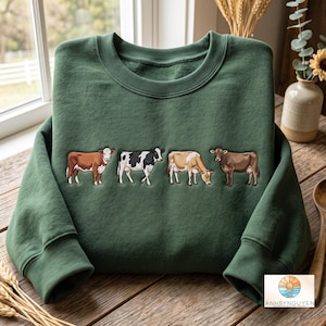 Könnte beinhalten: Ein waldgrüner Rundhals-Pullover mit einer horizontalen Reihe von vier Kühen in verschiedenen Farben und Mustern, darunter eine braun-weiße Kuh, eine schwarz-weiß gefleckte Kuh, eine hellbraune Kuh und eine einfarbig braune Kuh, die in einer Reihe stehen.