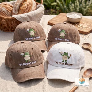 May include: Three brown and one white baseball cap featuring an embroidered green frog wearing an apron, holding a bowl and a wooden spoon, with the text "WE PROOF AT DAWN" printed in a clean font below the graphic on each of the four caps.
