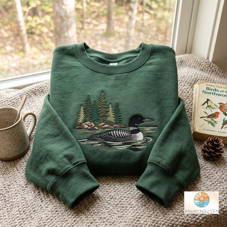 Puede incluir: Una sudadera de cuello redondo en color verde bosque con un dise&ntilde;o bordado de un colimbo nadando en el agua frente a un grupo de pinos. El bordado utiliza tonos negros, blancos y verdes para representar al ave y el paisaje forestal circundante.