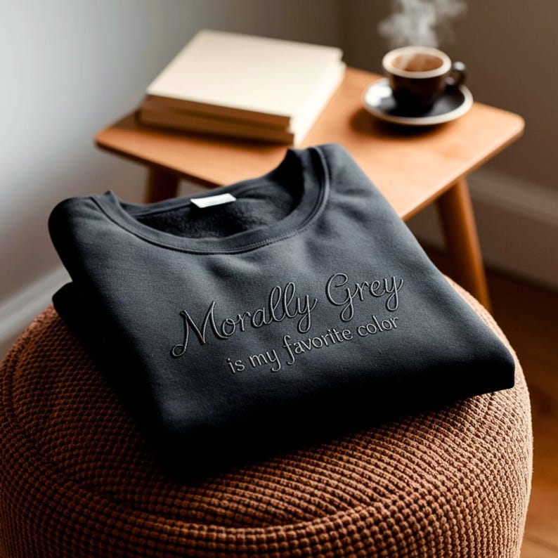 May include: A dark gray sweatshirt with the embroidered text "Morally Grey is my favorite color." The sweatshirt is folded on a brown woven ottoman. A small wooden table holds a stack of books and a cup of coffee.