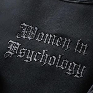May include: A black sweatshirt featuring the embroidered words "Women in Psychology" in a gothic style font. The text is a light grey colour, providing contrast against the dark fabric. The material of the sweatshirt appears soft.