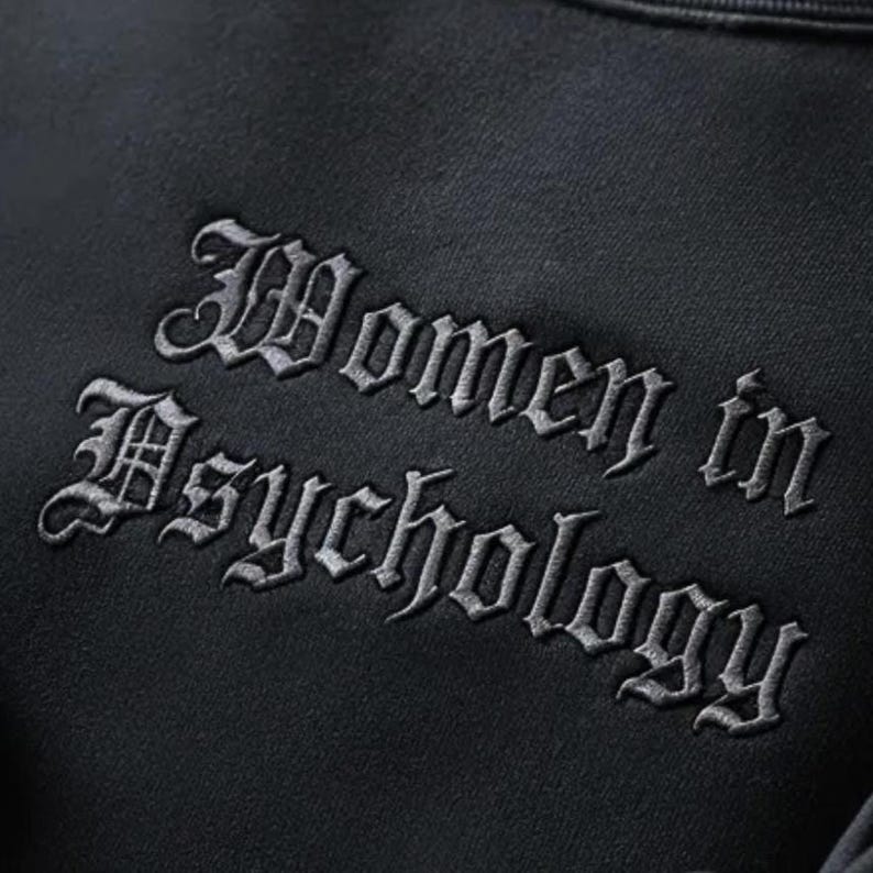 May include: A black sweatshirt featuring the embroidered words "Women in Psychology" in a gothic style font. The text is a light grey colour, providing contrast against the dark fabric. The material of the sweatshirt appears soft.