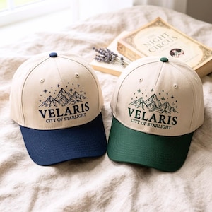 May include: Two baseball caps with the text "VELARIS CITY OF STARLIGHT". One cap has a navy blue brim and the other has a green brim. Both caps have a beige crown and a mountain and star design.