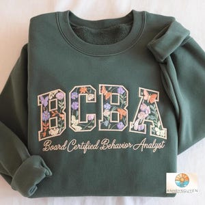 Op de afbeelding: Een donkergroene sweater met ronde hals en de geborduurde letters BCBA in een collegiaal lettertype. De letters zijn gevuld met bloem- en vlindermotieven in roze, paarse en oranje tinten. Daaronder is de tekst Board Certified Behavior Analyst geborduurd in een wit cursief lettertype.