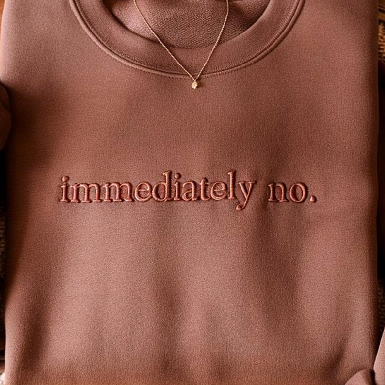 Peut inclure: Un sweat-shirt marron avec le texte brod&eacute; "immediately no." Un d&eacute;licat collier en or avec un petit pendentif est visible. Le sweat-shirt est de couleur unie et semble &ecirc;tre fait d'une mati&egrave;re douce.
