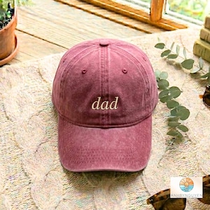 Puede incluir: Una gorra de béisbol color granate con una textura desgastada y lavada. La palabra dad está bordada en una fuente cursiva de color claro en el centro de la parte delantera de la gorra. La gorra presenta una corona estructurada, visera curva y costuras.