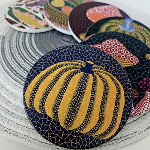 Puede incluir: Un juego de posavasos circulares de cerámica con motivos de calabazas punteadas en colores vibrantes como amarillo, azul, rojo y verde. Cada posavasos presenta un patrón intrincado de pequeños puntos y formas orgánicas muy detalladas.