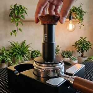 Peut inclure: Un moulin à café noir avec une poignée et un dessus en bois est utilisé. Le moulin est posé sur une base noire avec un porte-filtre en acier inoxydable. L'arrière-plan comprend des plantes et une ampoule.