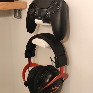 Op de afbeelding: Een zwarte gamecontroller en een zwarte en rode gamingheadset zijn aan een witte muur bevestigd. De koptelefoon heeft een rode accentkleur en het HyperX-logo. De controller is zwart.