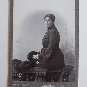Può includere: Una fotografia vintage in bianco e nero su cartoncino raffigurante una persona seduta di profilo, che indossa un indumento scuro a collo alto con maniche lunghe e una lunga collana. Il bordo inferiore riporta il testo cirillico Gr. Berman Minsk.