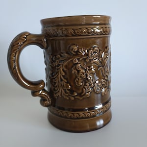 May include: A brown ceramic mug featuring a glossy finish and an intricate, raised relief pattern of scrolling leaves and vines. The mug has a thick, curved handle and decorative rope-like bands encircling both the top and bottom edges.