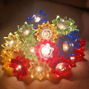 Op de afbeelding: Een lichtsnoer met decoratieve, doorschijnende stervormige kapjes in levendige kleuren rood, blauw, geel en groen. Elk lampje zit in een gefacetteerde, veelpuntige kunststof behuizing die oplicht wanneer de lampjes branden.