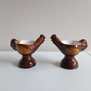Op de afbeelding: Twee bruine keramieken eierdopjes in de vorm van een haan. Elk dopje heeft een glanzende afwerking met een witte binnenkant voor een ei, een voetstuk en een gebeeldhouwde kop en staart. De hanen staan in tegengestelde richtingen.