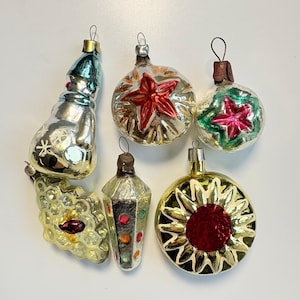 Könnte beinhalten: Eine Sammlung von sechs Vintage-Glasornamenten, bestehend aus einem silbernen Schneemann, einer Kugel mit rotem Stern, einer Kugel mit rosa Stern, einem strukturierten Cluster, einem hohen facettierten Ornament mit bunten Punkten und einer goldenen Sonne.