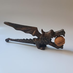 May include: A bronze-toned metal nutcracker shaped like a dragon with detailed wings and a scaled tail. The dragon's open jaws are holding a single brown, textured walnut. The lever mechanism is integrated into the dragon's wing and body design.