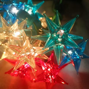 May include: A string of decorative star-shaped lights featuring translucent plastic covers in shades of red, blue, and clear white. Each star has multiple pointed rays and is illuminated from within, creating a glowing effect.