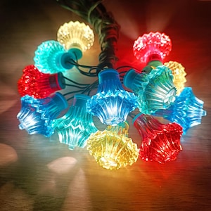 Könnte beinhalten: Eine dekorative LED-Lichterkette mit facettierten, glockenförmigen Kunststoffabdeckungen in den Farben Rot, Blau, Gelb und Türkis. Die Lichter sind durch ein dunkelgrünes Kabel verbunden und strahlen hell aus jedem der durchscheinenden Gehäuse.