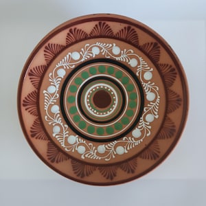 May include: A round ceramic plate with a terracotta base color featuring a dark brown scalloped border pattern. The center displays concentric circles of green dots, black rings, and a white vine motif with small white circular accents.