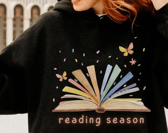 May include: A black hooded sweatshirt featuring a graphic of an open book with colorful pages fanning out, surrounded by small butterflies and confetti. The text "reading season" is printed in a light brown font below the book illustration.