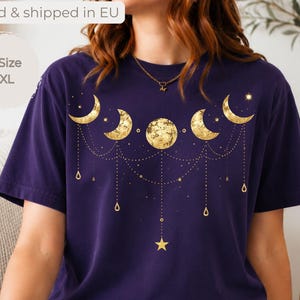 Op de afbeelding: Een donkerpaars t-shirt met korte mouwen en een gouden grafische print van maanfasen, inclusief sikkels en een volle maan, verbonden door fijne hangende kettinkjes en kleine sterren op de borst van het kledingstuk.