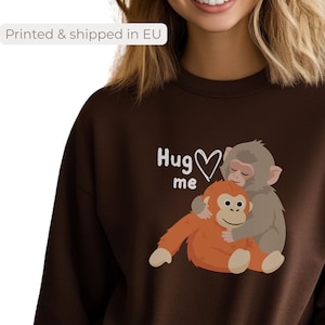 Könnte beinhalten: Ein braunes Sweatshirt mit einer Grafik von zwei Cartoon-Affen, die sich umarmen. Der Text "Hug me" steht über den Affen. Das Sweatshirt hat eine einfarbige braune Farbe.