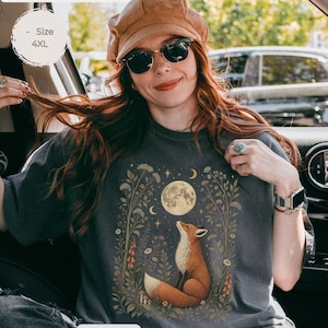 Può includere: Una t-shirt grigio antracite con una stampa grafica di una volpe seduta tra fiori selvatici e fogliame, che guarda verso una luna piena e stelle. La t-shirt ha un girocollo e maniche corte, con la parola pepper stampata in basso.