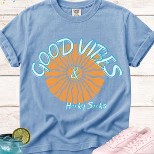 May include: Light blue Comfort Colors t-shirt with rolled sleeves. The shirt features a graphic of an orange sunburst with the words "GOOD VIBES & Hacky Sacks" in white and light blue. Pink sneakers are next to the shirt.