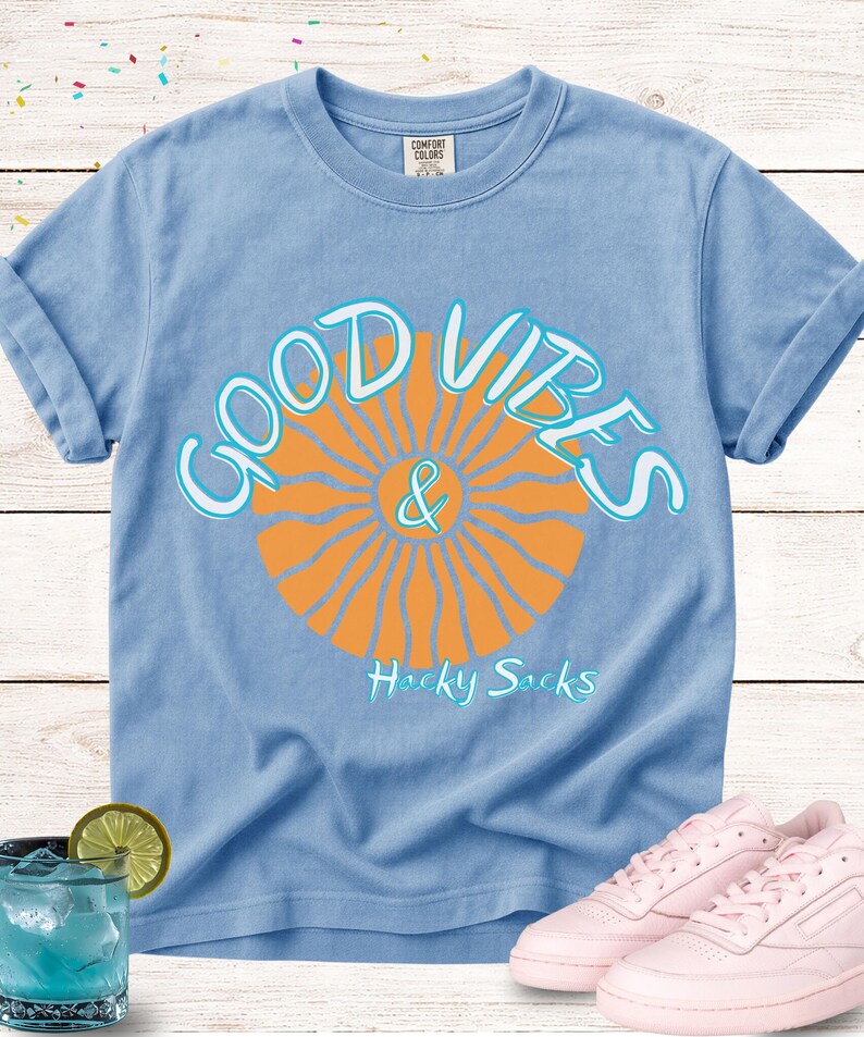 May include: Light blue Comfort Colors t-shirt with rolled sleeves. The shirt features a graphic of an orange sunburst with the words "GOOD VIBES & Hacky Sacks" in white and light blue. Pink sneakers are next to the shirt.