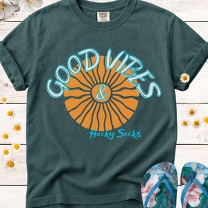 May include: Dark teal t-shirt with the words "GOOD VIBES & Hacky Sacks" in a retro font. The design features an orange sunburst graphic. A pair of floral flip-flops are at the bottom of the image.