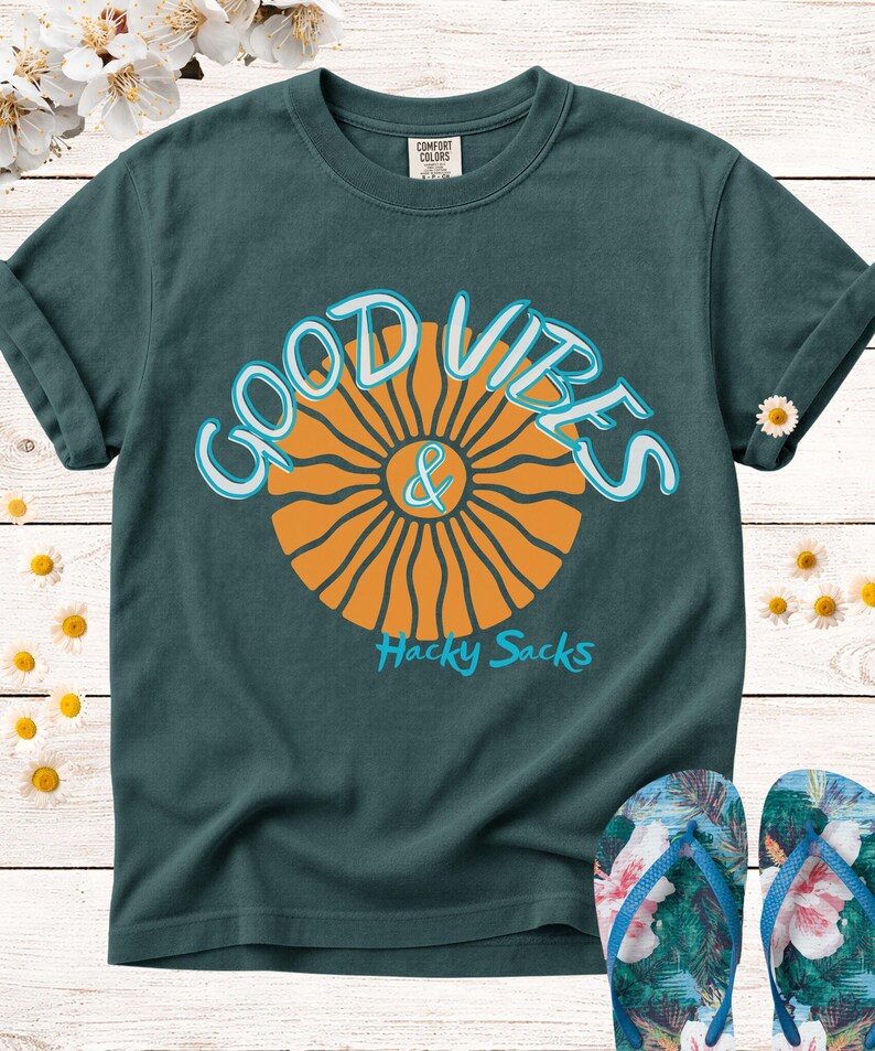 May include: Dark teal t-shirt with the words "GOOD VIBES & Hacky Sacks" in a retro font. The design features an orange sunburst graphic. A pair of floral flip-flops are at the bottom of the image.