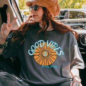 May include: A dark gray t-shirt with the words "GOOD VIBES & Hacky Sacks" in a blue and orange design. The shirt is worn with a brown corduroy hat and sunglasses.
