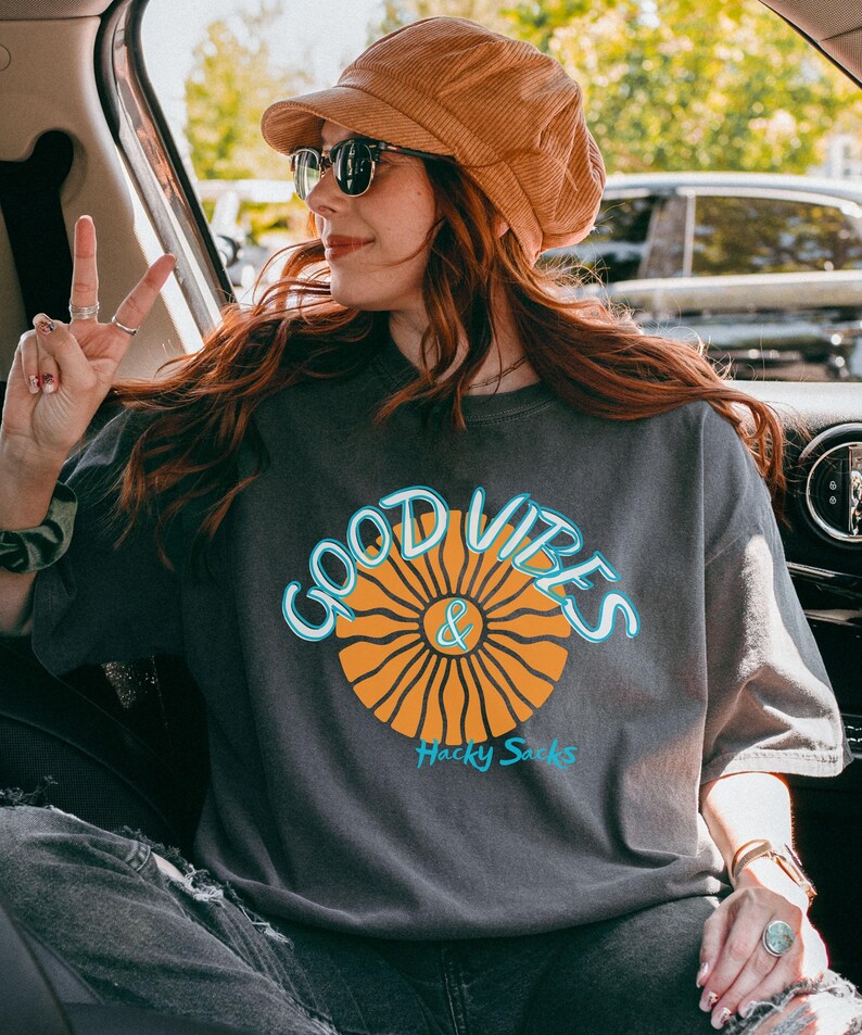 May include: A dark gray t-shirt with the words "GOOD VIBES & Hacky Sacks" in a blue and orange design. The shirt is worn with a brown corduroy hat and sunglasses.