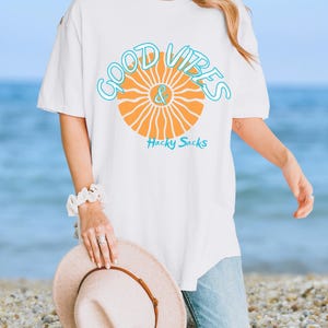 May include: White t-shirt with the words "Good Vibes & Hacky Sacks" in a turquoise and orange design. The shirt is a relaxed fit and is being held by a person on a beach.