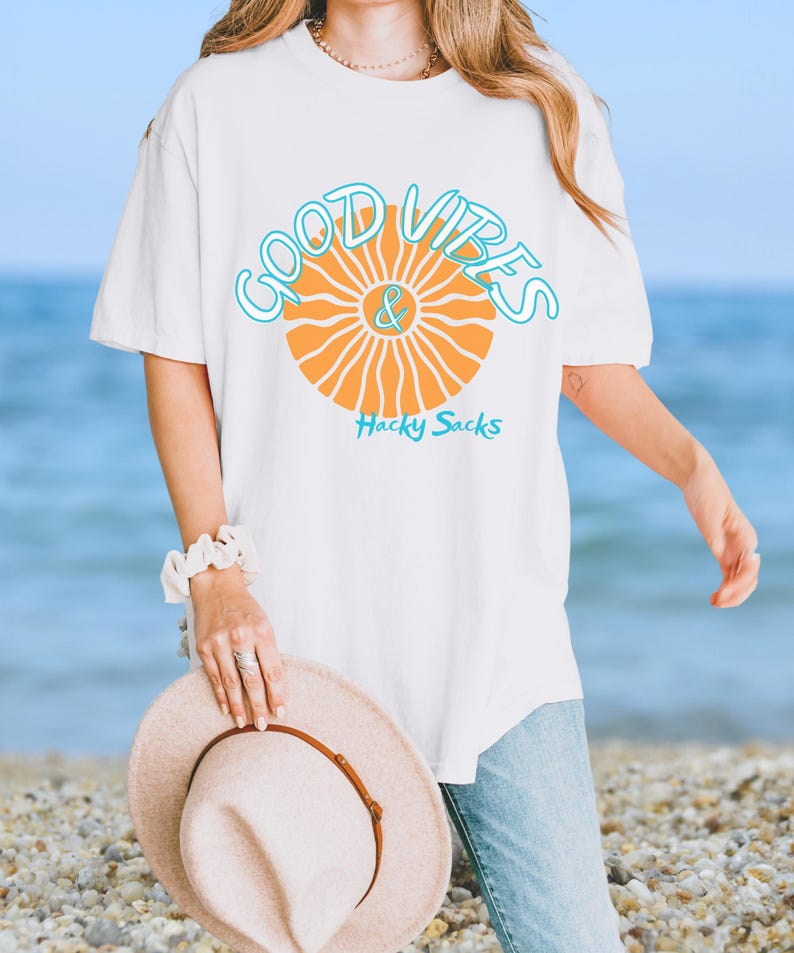 May include: White t-shirt with the words "Good Vibes & Hacky Sacks" in a turquoise and orange design. The shirt is a relaxed fit and is being held by a person on a beach.