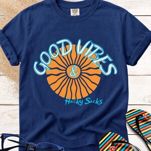 May include: Navy blue t-shirt with a graphic that says "GOOD VIBES & Hacky Sacks". The design features an orange sunburst and light blue lettering. A pair of blue glasses and colorful flip-flops are in the image.