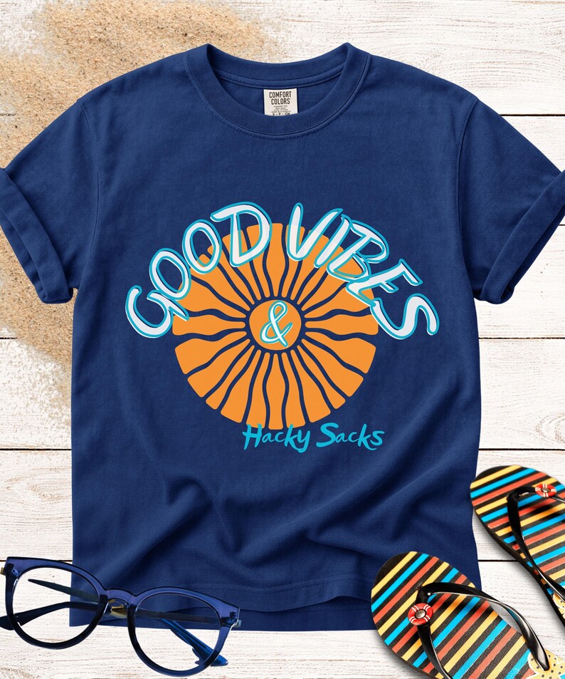 May include: Navy blue t-shirt with a graphic that says "GOOD VIBES & Hacky Sacks". The design features an orange sunburst and light blue lettering. A pair of blue glasses and colorful flip-flops are in the image.