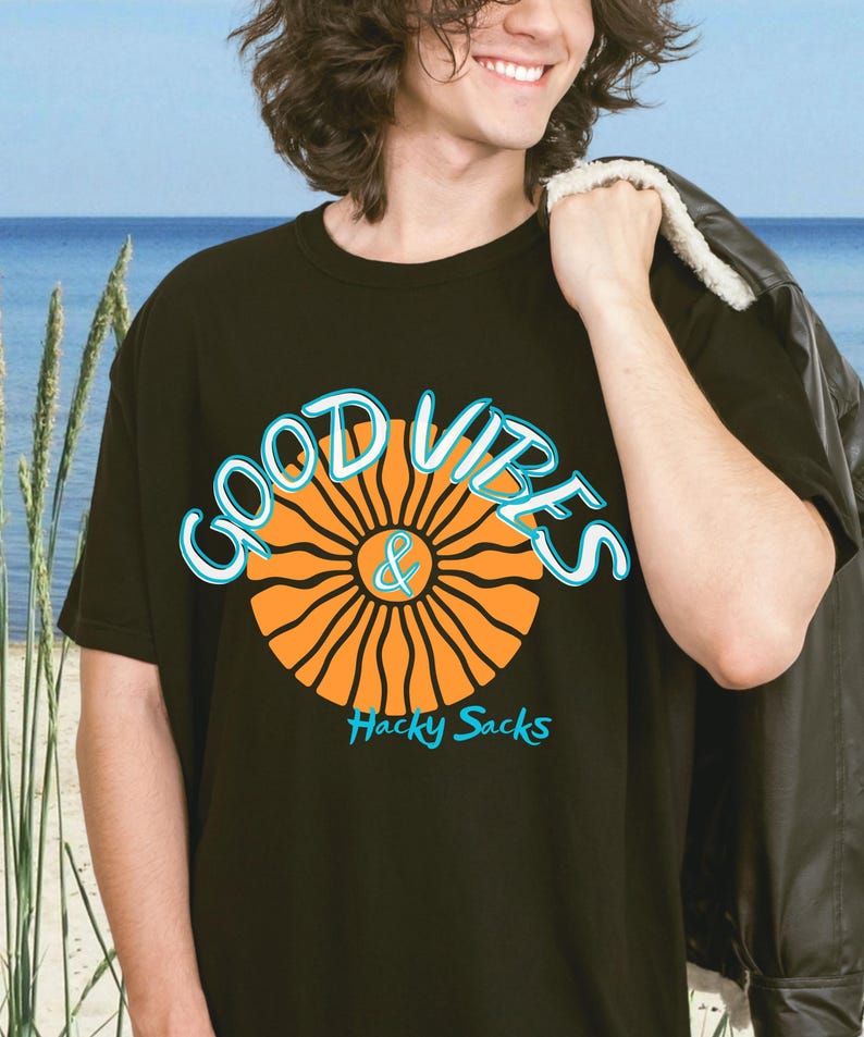 Young man smiling at the beach wearing a black retro graphic Comfort Colors t-shirt with colorful “Good Vibes & Hacky Sacks” design featuring an orange sun and turquoise lettering. Casual summer lifestyle shirt with beach and coastal aesthetic.
