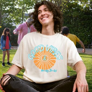 May include: Cream-colored t-shirt with the words "GOOD VIBES & Hacky Sacks" in a blue and orange design. The shirt is worn by a person sitting on the grass. The person is smiling. Other people are in the background.