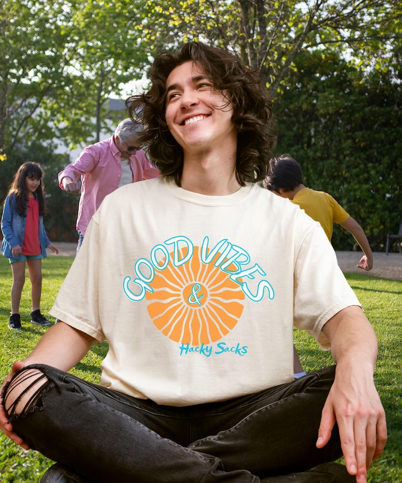 May include: Cream-colored t-shirt with the words "GOOD VIBES & Hacky Sacks" in a blue and orange design. The shirt is worn by a person sitting on the grass. The person is smiling. Other people are in the background.