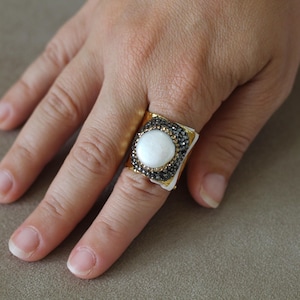 May include: A gold-toned ring with a square face featuring a large, round, white stone. The stone is surrounded by a border of small, dark gray stones. The ring is on a finger.