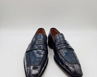 May include: A pair of navy blue leather loafers with a crocodile skin texture. The shoes have a slip-on design with a strap across the top and a square toe. The shoes are set against a white background.
