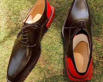 May include: Pair of black leather dress shoes with red accents on the heels. The shoes have a square toe design and lace-up closure. The shoes are displayed on a bed of green grass.