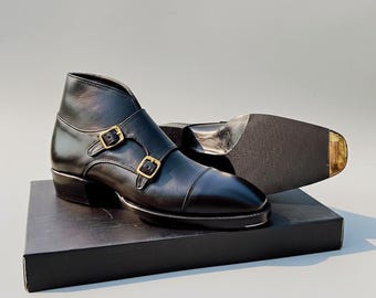 Puede incluir: Botines de cuero negro con hebillas dobles doradas. Los botines tienen un diseño de puntera y se exhiben sobre una caja negra. Se ve la suela de una bota, mostrando un patrón texturizado.
