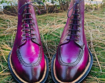 May include: A pair of burgundy leather boots with a high-top design. The boots have a lace-up closure with dark purple laces and gold-coloured eyelets. The soles have a black base with a tan-coloured trim. The boots are set on grass.