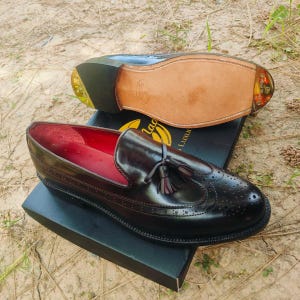 May include: Black leather loafers with decorative perforations and tassel accents. The shoes have a red interior and are displayed on a black box. The sole of one shoe is visible, showing a light brown leather sole.
