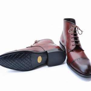 May include: Two brown leather boots with a classic design. The boots have a lace-up closure, a cap toe, and a black sole. The leather has a polished finish, and the boots are set against a white background.